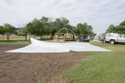 Several people drag a large white sheet towards a geodesic dome structure in a park area. 