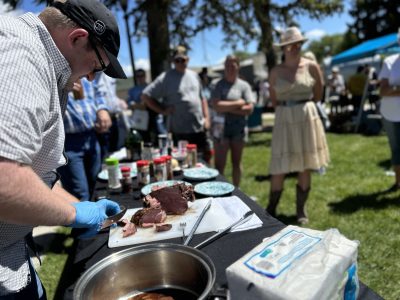 A man cuts up meat as people look on. The table is covered in different spice bottles and other cooking implements.