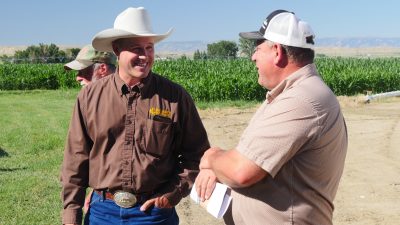 Two men talking near a field. One is wearing a cowboy hat, brown collared shirt, and belt buckle. The other is wearing a collared short sleeve and baseball cap.