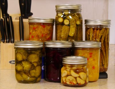 seven jars of canned goods, including pickles, pickled asparagus, and jam, sitting on a countertop with a knife block behind them