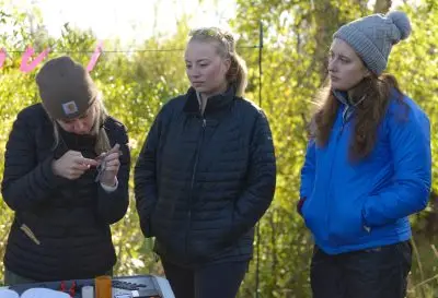 Two women look on as another woman holds a tool up to a small bird in her hand.