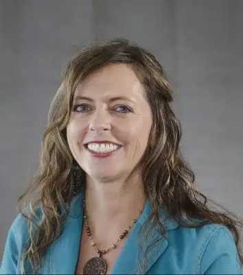 A woman with long wavy brown hair and blue eyes wearing a teal jacket and a large necklace.