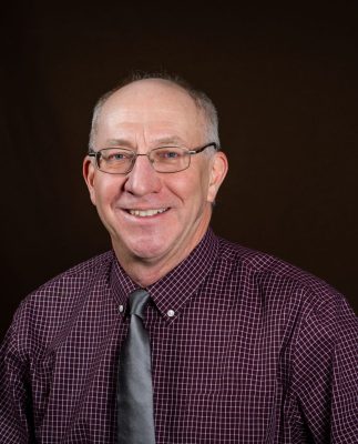 A white man with cropped gray hair wearing wire glasses, a maroon collared shirt, and a gray tie. 