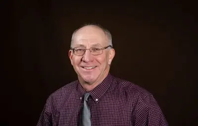A white man with cropped gray hair wearing wire glasses, a maroon collared shirt, and a gray tie.