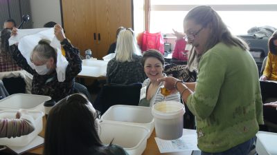 A woman stands holding a bucket and chatting with several women sitting at tables covered with large tubs.