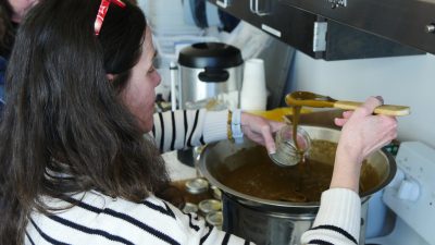 A woman uses a wooden spoon to ladle honey out of a large pot and into a mason jar.