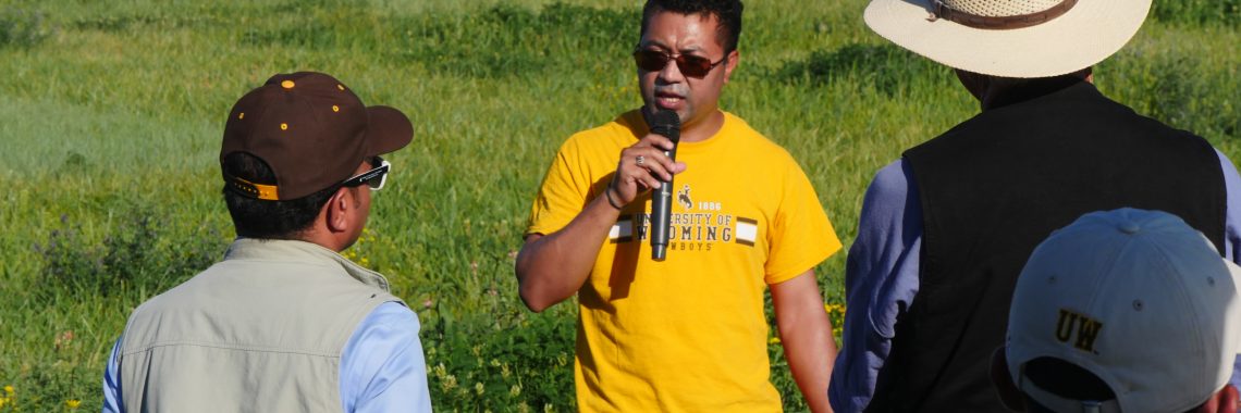 Participants listen to a young man with brown skin and short black hair wearing a UW Cowboys shirt speaking into a microphone in a field.