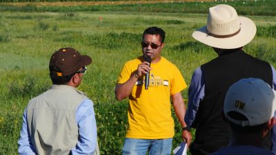 Participants listen to a young man with brown skin and short black hair wearing a UW Cowboys shirt speaking into a microphone in a field.