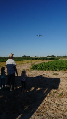 A man and two kids watch a drone flying over a field. 