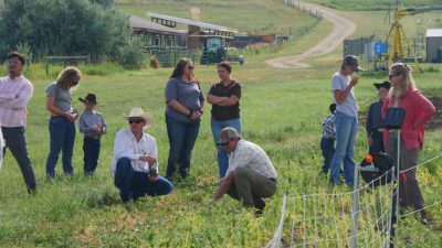 Five women, three young boys in cowboy hats, and three men in a field. Two of the men are crouching down amidst some weeds. There are fences, tractors, and agricultural buildings in the background, as well as a dirt road.