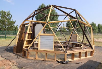 geodesic dome frame with dark brown wood and yellow wood beams, internal raised beds with irrigation system  and small pile of black tubing in front. The dome is surrounded by chainlink fence with trees on the other side of the fence