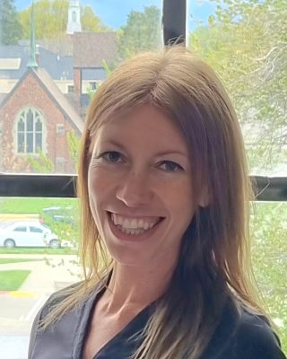 portrait of smiling woman with short brown hair and black shirt standing in front of a window 