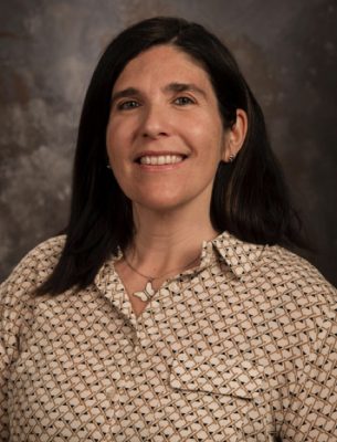 A woman with straight black hair a little longer than her shoulders and pale skin wearing a checked collared shirt and a butterfly necklace. 