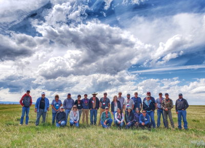 24 people, most of them wearing name tags on red lanyards stand in a grassy pasture with blue sky and clouds overhead