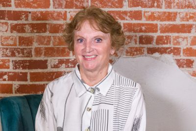 An older woman with light skin, blue eyes, and short light brown wavy hair. She is wearing a white button up with abstract line patterns. 