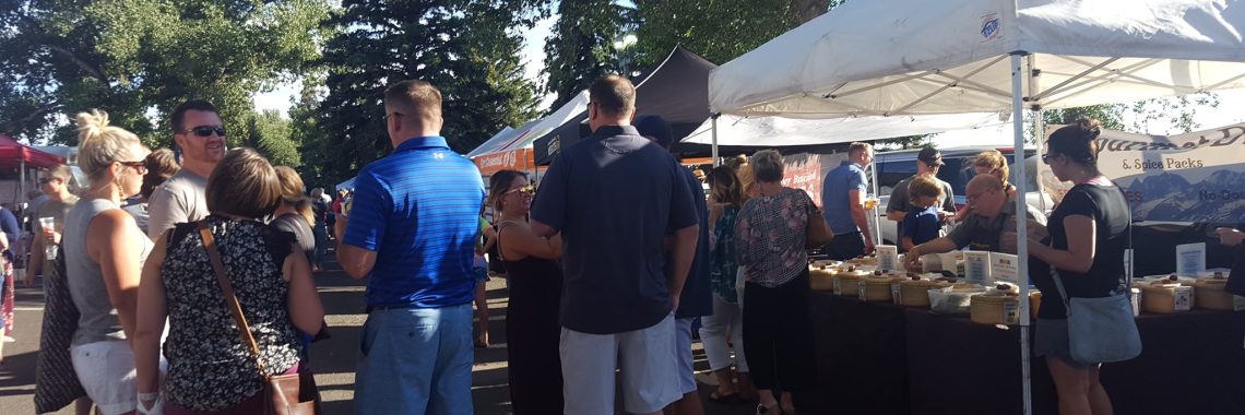 group of people chat at a crowded farmers' market in a parking lot bordered with trees next to a white vendor tent on a sunny day