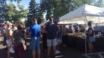 group of people chat at a crowded farmers' market in a parking lot bordered with trees next to a white vendor tent on a sunny day