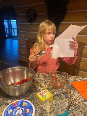 young girl with blond hair and bangs wearing pink sweatshirt holds paper recipe in one hand and spoon in the other while standing in front of a countertop with a metal bowl of strawberries, glass measuring cup, lemon Jello packet, and Cool Whip container