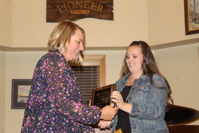 A woman with bobbed blonde hair accepts a plaque from another woman with long brown hair. 