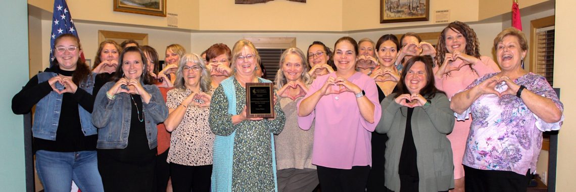 A group of about 20 middle aged white women in semi-formal wear, all making heart shapes with their hands. In the middle, a woman holds up a plaque.