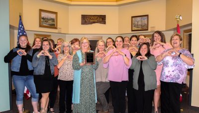 A group of about 20 middle aged white women in semi-formal wear, all making heart shapes with their hands. In the middle, a woman holds up a plaque.