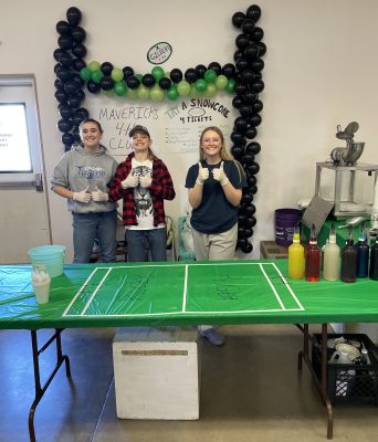 three smiling teenagers wearing disposable gloves give double thumbs up to the camera while standing under a balloon arch attached to the wall and a sign advertising snow cones made by the Mavericks 4-H club. Syrups and a snow cone machine are lined up on the tables in front and to the side of them