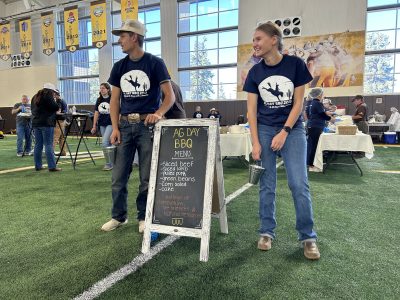 Two smiling young people in Ag Day BBQ t-shirts and jeans stand next to a sign that reads "Ag Day BBQ. Menu. Silced beef, sliced lamb, pulled pork, green beans, corn salad, cake. College of agriculture, life sciences and natural resources." 