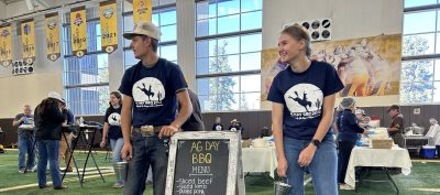 Two smiling young people in Ag Day BBQ t-shirts and jeans stand next to a sign that reads "Ag Day BBQ. Menu. Silced beef, sliced lamb, pulled pork, green beans, corn salad, cake. College of agriculture, life sciences and natural resources."