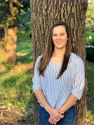 A white woman with long straight brown hair leans against a tree.