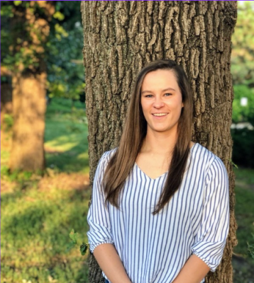 A white woman with long straight brown hair leans against a tree.