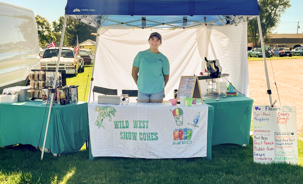 smiling teenage girl in a turquoise shirt t-shirt and shorts stands under an open-sided tent behind a table draped with a banner labeled Wild West Snow Cones. To her left is a snow cone machine and a sign with list of flavors; to her right is a table with a display of insulated mugs.