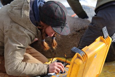 A woman dressed in warm clothes leans down to examine something within a yellow plastic case. 