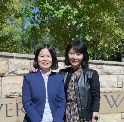 Two Chinese women standing next to each other. They both have bobbed black straight hair and light skin. The woman on the left is wearing a blue suit and the woman on the right is wearing a dress and black jacket.