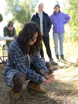 A woman in plaid kneeling down to release a dark brown bird with white spots while several people look on. 