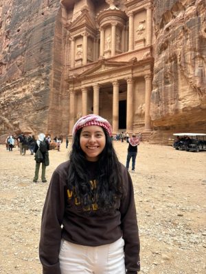 A young woman with long straight black hair and light brown skin wearing a hat. A temple with carved columns is in the back.