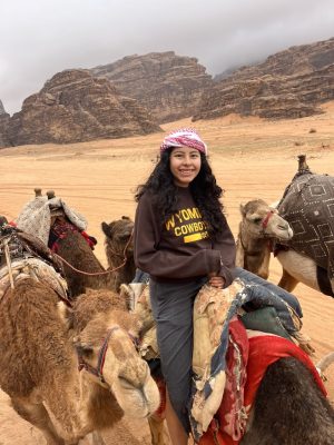 A young woman wearing a Wyoming Cowboys shirt in a desert on a camel with several camels around her. There are jagged rock formations in the background.