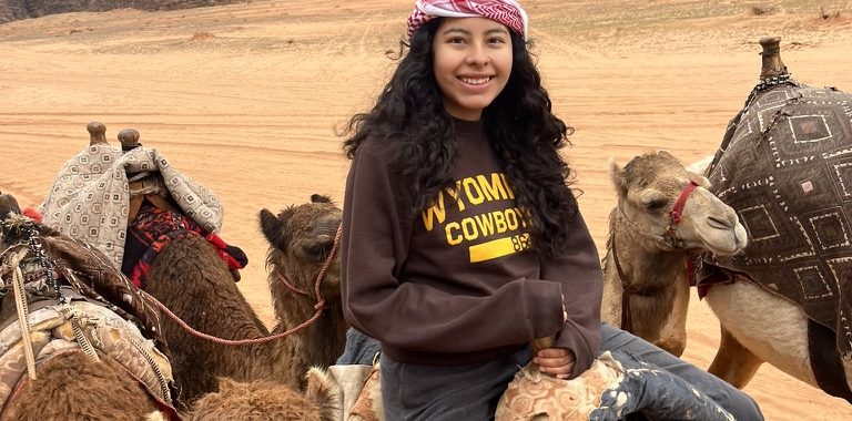 A young woman wearing a Wyoming Cowboys shirt in a desert on a camel with several camels around her. There are jagged rock formations in the background.