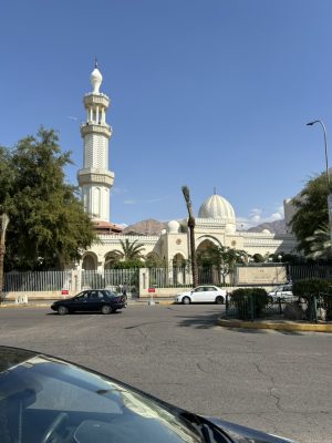 A white building with a hexagonal tower and two domes, viewed from across a parking lot. Several palm trees are in front of it.