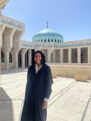 A young woman wearing a black robe in a courtyard, which is made of white stone. In the background is a blue dome and there's blue writing above the columns holding up the walls of the courtyard. 