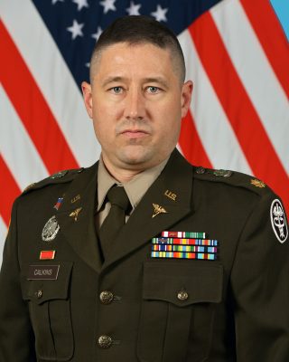 A white man with short black hair and blue eyes wearing a military uniform in front of an American flag.