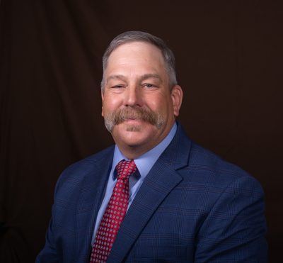 A man with short gray hair and a large mustache wearing a blue suit and red tie. 