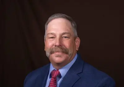 A man with short gray hair and a large mustache wearing a blue suit and red tie.
