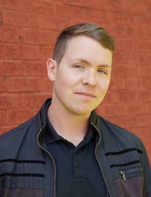 A young man with pale skin and coppery short straight hair. He's wearing a black shirt and black jacket. 