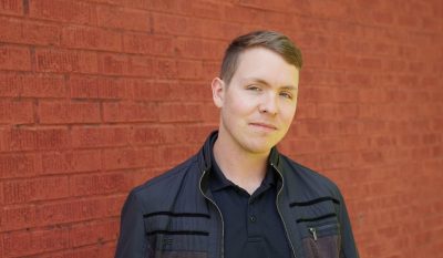 A young man with pale skin and coppery short straight hair. He's wearing a black shirt and black jacket.