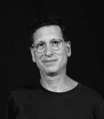 A black and white picture of an older man with wire glasses and short straight hair wearing a t-shirt.