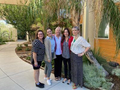 four smiling women stand together for a group photo on a concrete pathway in front of a garden bordering a building