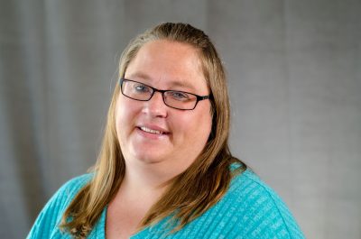 A woman with light skin and shoulder length straight dark blonde hair. She is wearing square glasses and a bright teal shirt.