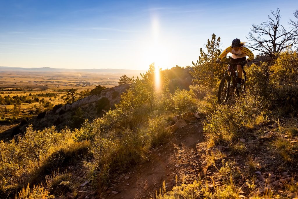 mountain biker wearing a yellow long-sleeve jersey rides down a rocky slope toward the camera. A valley with small outcroppings of trees spreads out toward the mountains behind him.