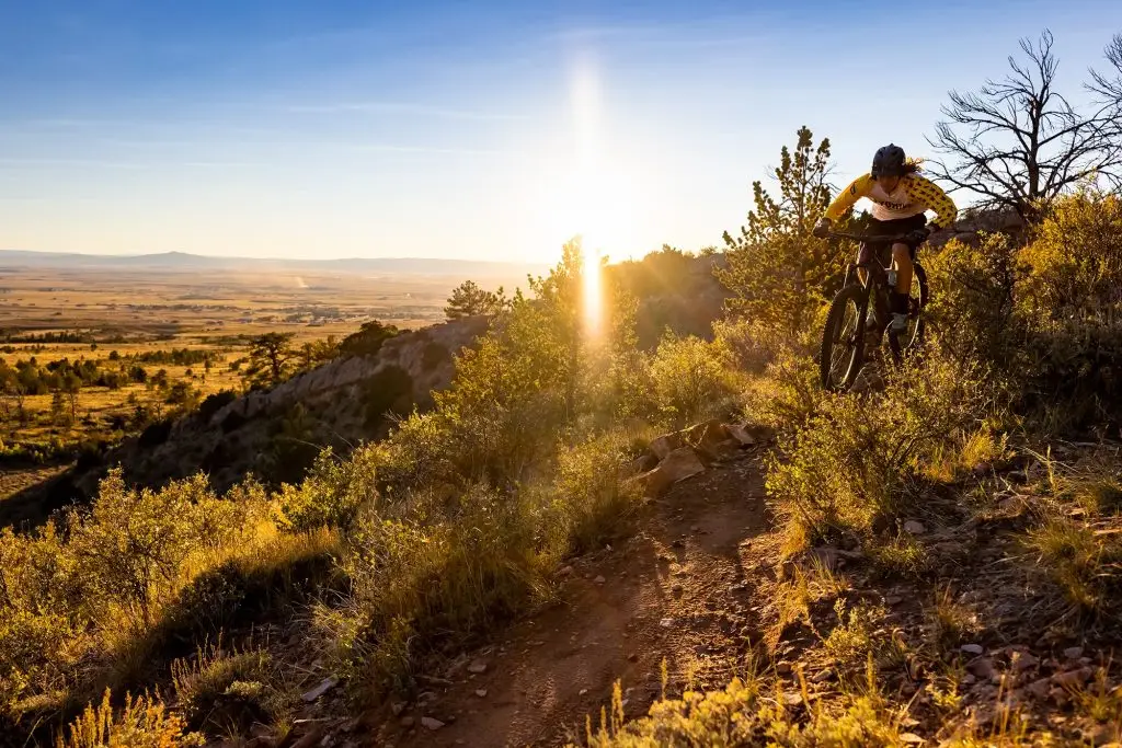 mountain biker wearing a yellow long-sleeve jersey rides down a rocky slope toward the camera. A valley with small outcroppings of trees spreads out toward the mountains behind him.