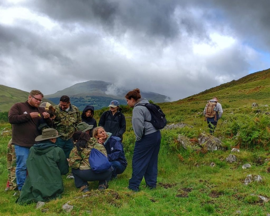 group of people, some wearing camouflage jackets, bend down to examine plants in a lush green meadow under a dramatic cloudy sky. A man wearing a brown quarter zip and jeans holds a piece of plant material, showing it to the others in the group